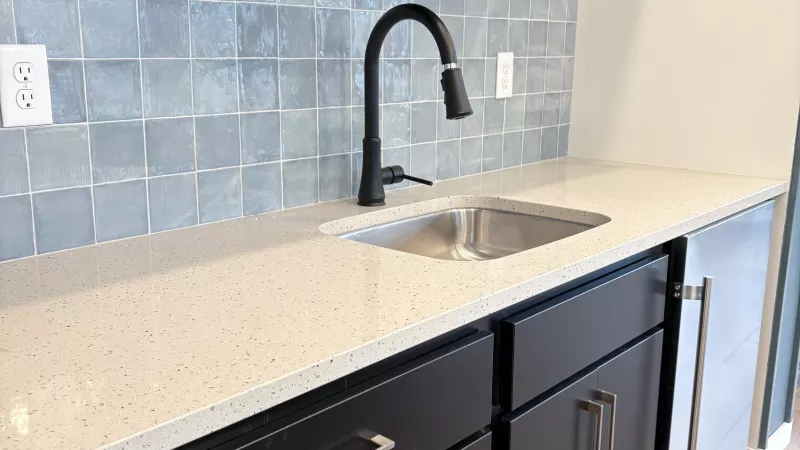 Modern kitchenette with a speckled light countertop, black cabinets, matte black faucet, and glossy blue tile backsplash beneath two floating shelves.