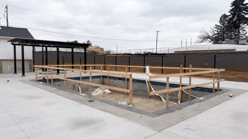 Outdoor pool under construction with wooden safety railings, surrounded by new concrete patio and fenced community grounds.