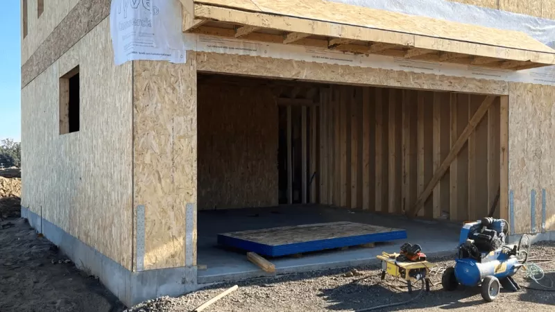 Exterior corner of a building with wood sheathing and a framed overhang above a large opening, accompanied by construction tools on the ground.