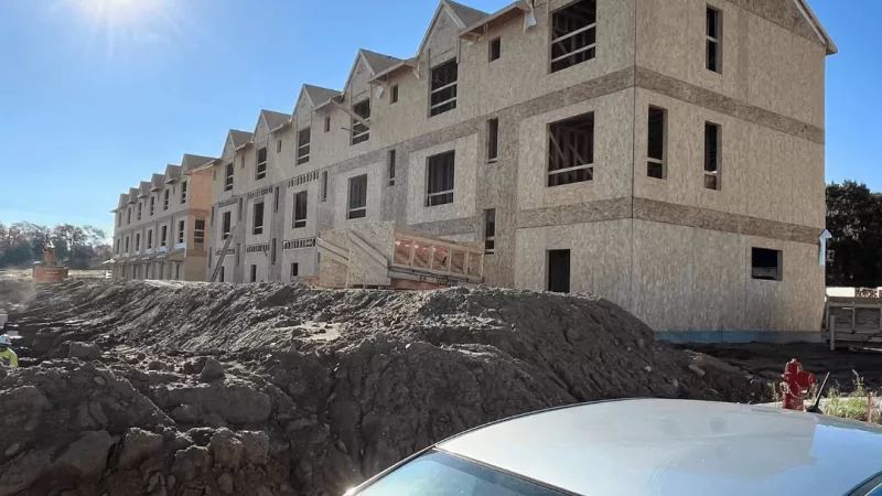 A row of multi-story townhomes fully framed with wood sheathing sitting behind a dirt mound under a bright sunny sky.