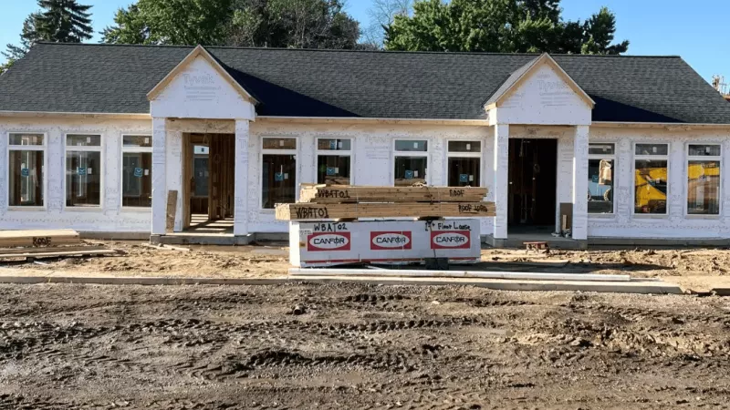 Exterior view of a single-story building under construction with installed windows, Tyvek wrap, and stacked lumber in the foreground.
