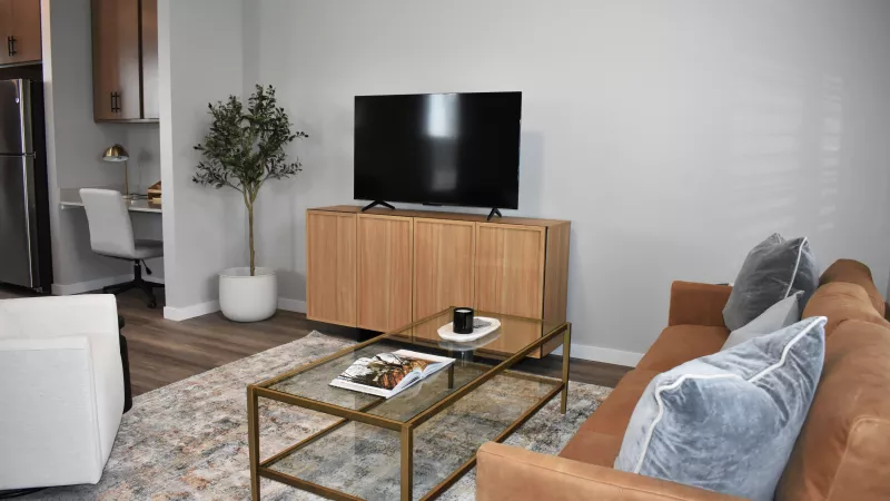 Stylish living room featuring a brown couch, glass coffee table, flat-screen TV, and a glimpse of a kitchen.