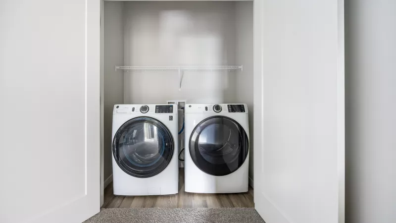 Two white front-loading washer and dryer machines in a bright, minimalist closet with a shelf above.