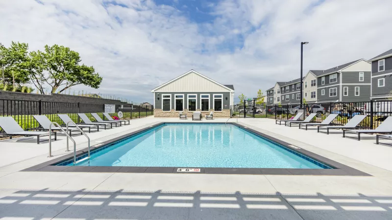 Outdoor swimming pool with lounge chairs, a clubhouse, and apartment buildings under a partly cloudy sky.