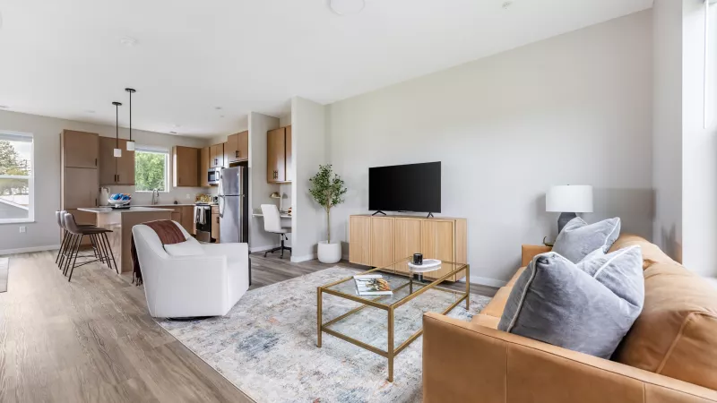 Bright, modern open-concept living room with a brown leather sofa, light wood media console, and an adjacent kitchen area.