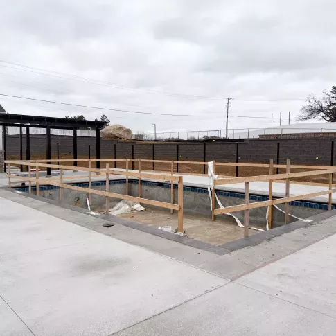 Outdoor pool under construction with wooden safety railings, surrounded by new concrete patio and fenced community grounds.