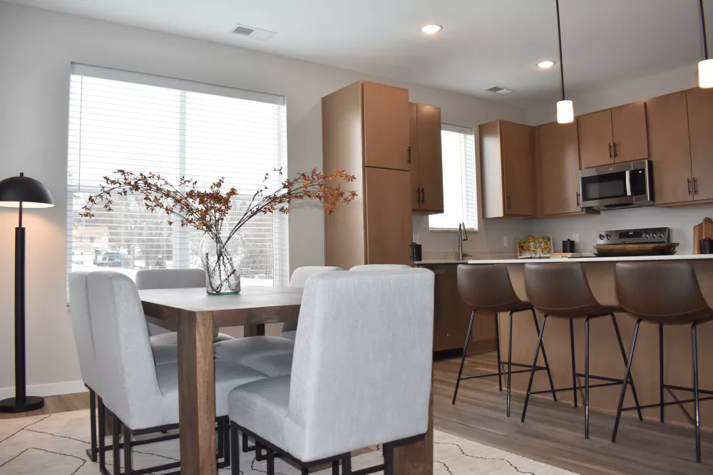 Modern open-concept dining area with wood table, grey chairs, and a kitchen featuring wood cabinets and island with brown stools.