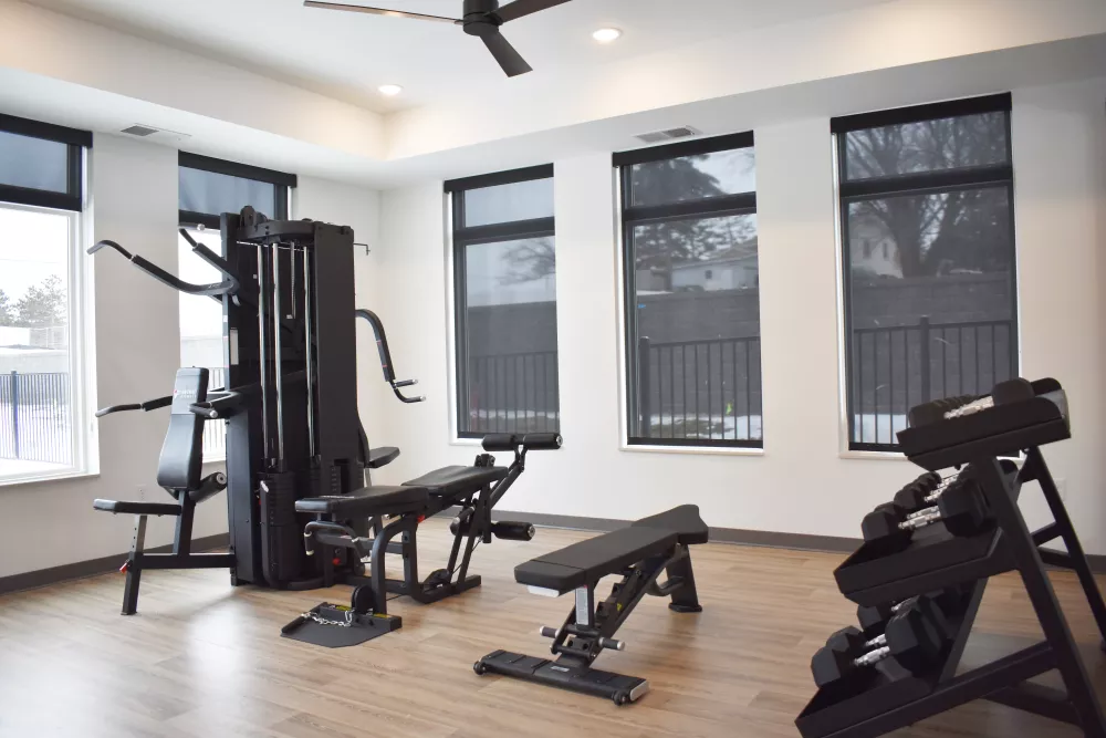 Well-lit modern gym with a multi-station machine, weight benches, and dumbbells on a wooden floor, white walls, and dark blinds.